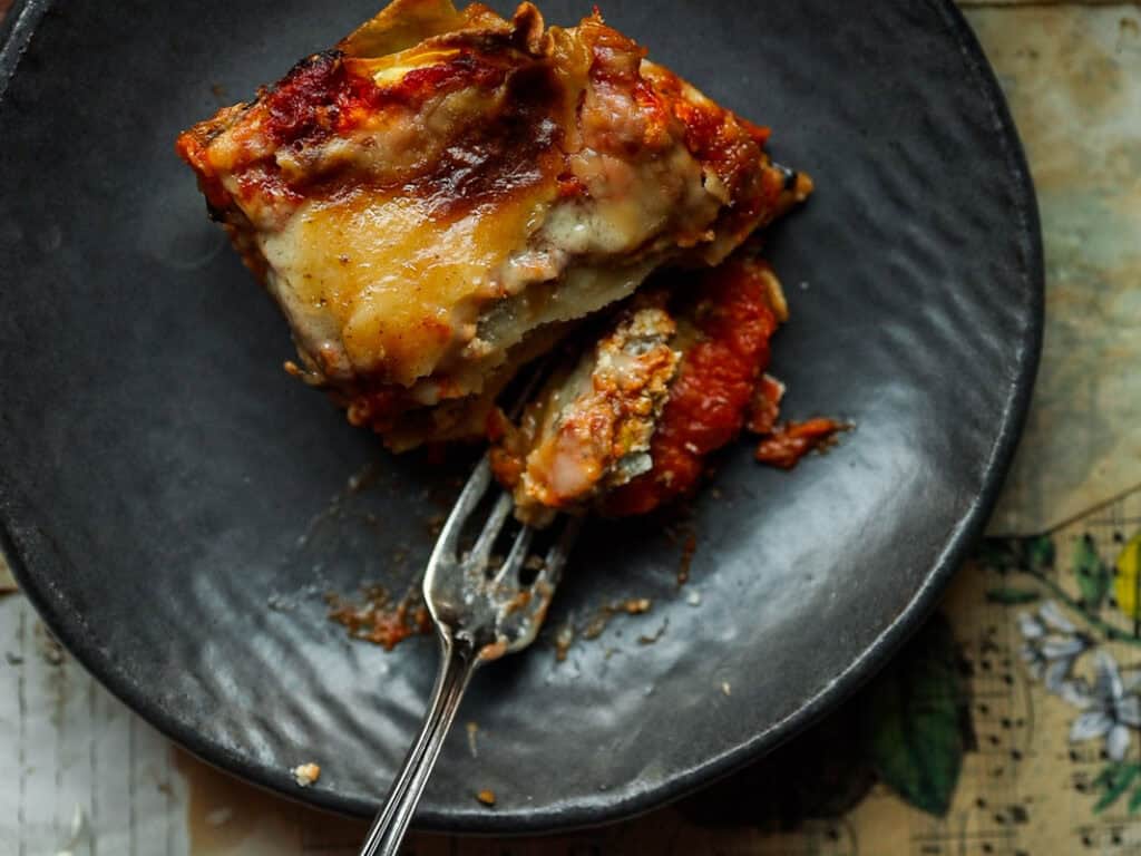 A slice of vegan lasagna is on a dark plate, with a fork partially inserted into it. The lasagna is layered with visible cheese, sauce, and pasta, with a slightly crispy top. It sits on a textured tabletop.